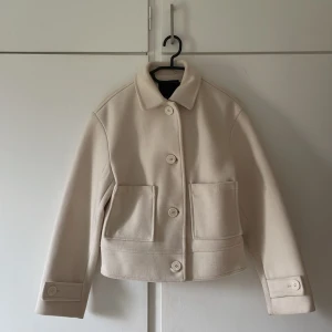 Beige ulljacka med stora knappar - Snygg beige jacka i ull med klassisk krage och stora knappar framtill. Jackan har två rejäla fickor och raka ärmar med knappdetalj vid mudden. Perfekt för dig som gillar stilren och minimalistisk look.