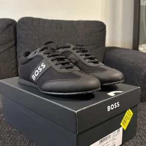 Svarta  BOSS skor  - Helt nya aldrig använt beställda från Boozt. Stilrena svarta sneakers från BOSS med tydlig logga på sidan. Skorna har en kombination av textil och syntetmaterial, snörning och platt sula. Perfekta för dig som gillar en clean och sportig look med diskreta detaljer. 