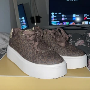 Bruna Michael Kors sneakers med platåsula - Snygga bruna sneakers från Michael Kors med monogrammönster och detaljer i guld. Skorna har en rejäl vit platåsula och snörning framtill. Ovandelen är i textil med guldiga detaljer på häl och plös, samt MK-logga bak. Perfekta för dig som vill sticka ut.
