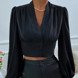 Svart croppad blus med knappar - Snygg svart croppad blus med djup v-ringning och långa puffärmar. Blusen har bred midjedel med dekorativa knappar framtill.