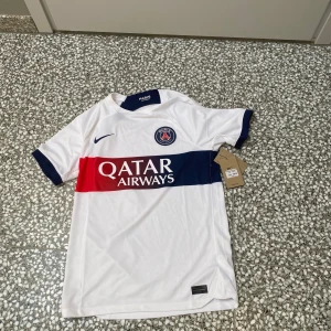 PSG vit matchtröja med blå och röd detalj - Säljer en vit Paris Saint-Germain fotbollströja från Nike med blå och röd rand över bröstet och Qatar Airways-tryck. PSG-logga och Nike-märke på framsidan, blå krage och ärmslut. Perfekt för fans!