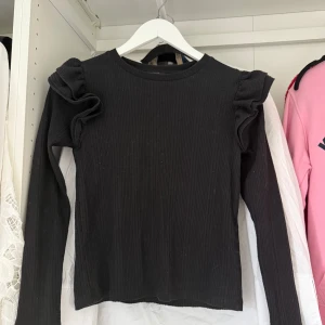 Svart ribbad topp med volangärmar från Zara - Säljer en svart långärmad topp från Zara med ribbad struktur och volangdetaljer på axlarna. Tröjan har en rund halsringning och är figurnära i modellen. Perfekt för dig som gillar stilrena plagg med en twist.