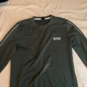 Mörkgrå långärmad tröja från Hugo Boss - Säljer en stilren mörkgrå långärmad tröja från Hugo Boss med vit logga på bröstet och diskreta vita ränder längs ärmarna. Perfekt för en clean och modern look.