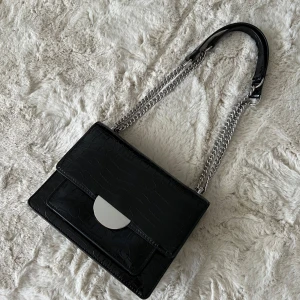 Svart axelväska med kedja och krokodilstruktur - Säljer en svart axelväska med snygg krokodilpräglad yta och silverfärgad kedjerem. 