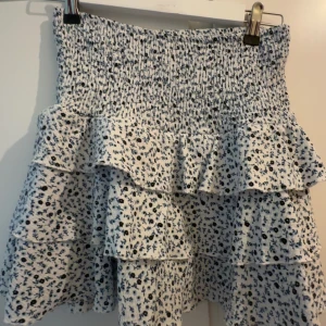 Vit blommig volangkjol - Supersöt vit kjol med blått blommönster och flera volanger. Kjolen har smock i midjan för en bekväm passform och är perfekt för varma dagar. Den är kort och har en luftig känsla.