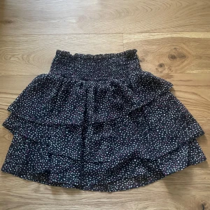 Svart volangkjol med småmönster - Supersöt svart kjol med volanger och små vita och rosa prickar. Kjolen har en smockad midja som gör den bekväm och sitter snyggt. Perfekt att styla med en enkel topp eller hoodie.