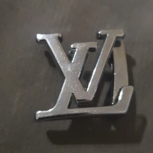 Silverfärgat Louis Vuitton bältesspänne - Säljer ett stilrent bältesspänne från Louis Vuitton i silverfärg. Spännet har den klassiska LV-loggan och är tillverkat i metall med märkning 835. Perfekt för att ge din outfit en exklusiv touch.