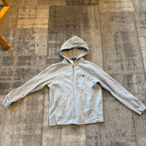 Ljusgrå hoodie från Polo Ralph Lauren - Ljusgrå hoodie med dragkedja från Polo Ralph Lauren. Tröjan har långa ärmar, huva och två fickor framtill. Klassisk liten broderad logga på bröstet. Perfekt för en avslappnad stil.