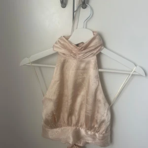 Beige halterneck topp i satin - Säljer en beige halterneck topp i glansigt satinmaterial med hög krage och knytning i ryggen. Toppen har en elegant och stilren look, perfekt för att lyfta vilken outfit som helst.