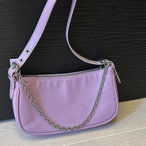 Ljuslila axelväska med kedjedetalj - Säljer en ljuslila axelväska i syntetmaterial med krokodilpräglad yta. Väskan har en silverfärgad kedja framtill och justerbart axelband. Stängs med dragkedja upptill och har ett stilrent, modernt utseende.