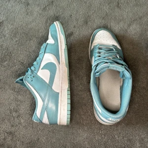 Nike Dunk sneakers i blått och vitt - Snygga Nike Dunk sneakers i ljusblått och vitt med klassisk design. Skorna har snörning, rund tå och platt sula. Perfekta för dig som vill ha en stilren och sportig look. Ikonisk Nike-logga på sidan och mjuk vaddering för extra komfort.