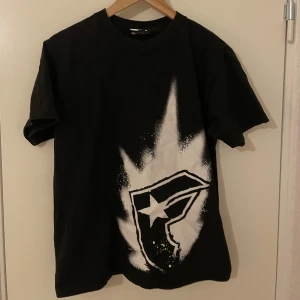 Svart t-shirt från Famous Stars and Straps - Svart t-shirt från Famous Stars and Straps med stort vitt sprayat logotryck framtill och textdetalj baktill. Klassisk passform och rund halsringning. Perfekt för dig som gillar streetwear och vill sticka ut med en unik design.