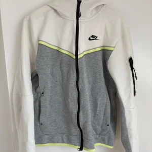 Sällsynt nike tech fleece - Sällsynt Nike tech fleece med dragkedja i vitt och grått med svarta detaljer och en tunn gul linje. Koftan har huva, lång ärm och fickor framtill samt en ficka med dragkedja på ena ärmen. Perfekt för en sportig look.