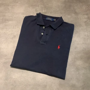 Mörkblå pikétröja från Polo Ralph Lauren - Säljer en klassisk mörkblå pikétröja från Polo Ralph Lauren med röd broderad logga på bröstet. Tröjan har korta ärmar, krage och knappar framtill. Perfekt för en stilren och avslappnad look. Tröjan har en defekt som du ser på bild 5, det är inte ett hål! Bara att höra av sig vid funderingar eller frågor! Priset är inte hugget i sten!