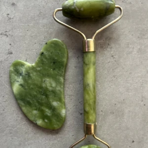 Grön jade ansiktsroller och gua sha - Säljer ett set med ansiktsroller och gua sha i grön jade. Rollern har två olika storlekar på stenarna och metallfästen i guldton. Perfekt för ansiktsmassage och hudvård. Gua sha-stenen är hjärtformad och matchar rollern i färg och material. Aldrig använda!