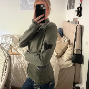 Olivgrön kavaj med nitar från Two Wings - Säljer en olivgrön kavaj från Two Wings med snygga nitar på krage och fickor. Kavajen har två knappar framtill, lång ärm och dekorativ patch på ena ärmen. Perfekt för dig som vill sticka ut lite extra.