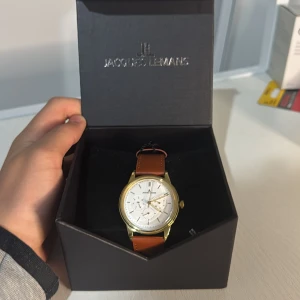 Jacques Lemans herrklocka med brunt skinnarmband - Stilren herrklocka från Jacques Lemans med vitt urtavla, guldigt boett och brunt skinnarmband. Klockan har flera små urtavlor och klassisk design. Perfekt accessoar för dig som gillar tidlös stil.