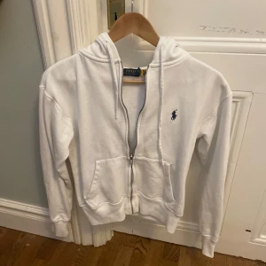 Vit hoodie med dragkedja från Polo Ralph Lauren - Säljer en klassisk vit hoodie från Polo Ralph Lauren med dragkedja och huva. Tröjan har två fickor framtill och den ikoniska broderade loggan på bröstet. Perfekt för en avslappnad stil.