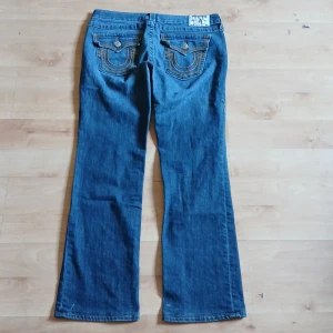 Blå jeansbyxor från True Religion - Säljer ett par klassiska blå jeansbyxor från True Religion med snygga kontrastsömmar och dekorativa ficklock bak. Byxorna har normal passform och bootcut-stil. Perfekta för dig som gillar tidlös denim med detaljer.