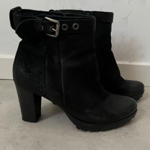 Svarta boots med klack och spänne - Säljer ett par svarta boots med hög klack och grov sula. Skorna har ett dekorativt spänne och dragkedja på sidan för enkel påtagning. Materialet är en mix av skinn och syntet med en matt finish. Perfekta för att lyfta din outfit.