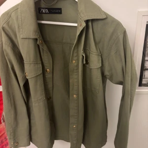 Olivgrön jeansjacka  från Zara - Säljer en olivgrön jeansjacka från Zara med klassisk krage och bröstficka. Jackan har guldiga knappar framtill och på fickorna. Perfekt att slänga över en t-shirt för en avslappnad look.