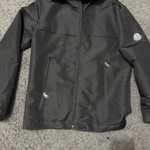 Svart vindjacka från Moncler - Säljer en svart vindjacka från Moncler med dragkedja och två snedställda fickor framtill. Jackan har Moncler-logga på ärmen och är tillverkad i ett slätt syntetmaterial. Perfekt för dig som gillar stilren och sportig look. 