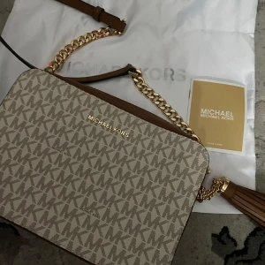 Beige och brun axelväska från Michael Kors - Snygg axelväska från Michael Kors i beige och brunt med MK-logga över hela väskan. Väskan har guldfärgade detaljer, kedjerem och tofs. Perfekt storlek för det viktigaste och har dragkedja upptill.