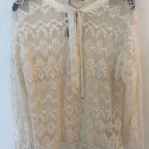 Vila vit blus, storlek M, helt ny med prislapp - En vit spetsblus med långa ärmar och en rund halsringning. Blusen har en snörning i midjan och en liten gulddetalj vid snörningen. Blusen är genomskinlig och har ett fint spetsmönster.  Helt ny med prislapp  Strl M