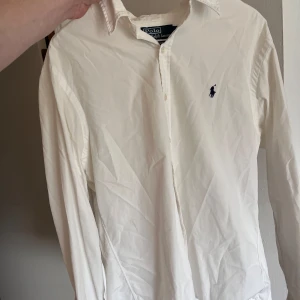 Vit skjorta från Polo Ralph Lauren - Klassisk vit skjorta från Polo Ralph Lauren med broderad marinblå logga på bröstet. Skjortan har lång ärm, knappar framtill och krage. Perfekt till dig som gillar stilrena och tidlösa plagg. Inga fläckar eller skador. Kom privat för mer info!