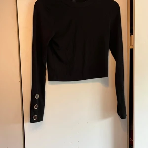 Svart croppad långärmad topp från Gina Tricot - Säljer en svart croppad långärmad topp från Gina Tricot. Toppen är ribbad och har tre stora knappar som detalj på ena ärmen. Perfekt till många olika outfits och enkel att matcha. Finns en liten fläck på ryggen (se sista bilden) därav priset, kanske någon som har super knep på att få bort fläckar som isf fyndar en super snygg topp