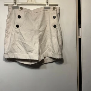 Beiga shorts med guld knappar - Stilrena beiga shorts från Escada med hög midja och dekorativa guldiga knappar framtill. Shortsen har uppvikta benslut och är tillverkade i ett bomullsliknande material. Perfekta för sommardagar när du vill ha en klassisk look. Diskuterbart pris