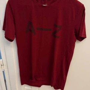 Vinröd t-shirt från A–Z - Säljer en vinröd t-shirt från A–Z med svart tryck på bröstet. T-shirten har rund halsringning och är tillverkad i mjuk bomull. Perfekt för en avslappnad sportig stil.