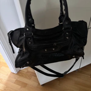 Svart axelväska med dragkedja - Säljer en svart axelväska i skinn med både handtag och axelrem. Väskan har flera nitar och detaljer samt en dragkedja framtill. .