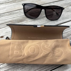 Svarta solglasögon från Hugo Boss - Stilrena svarta solglasögon från Hugo Boss med klassisk wayfarer-form. Glasögonen har mörka linser och diskret Hugo Boss-logga på skalmen. Kommer med originalfodral i svart med BOSS-tryck och beige putsduk.