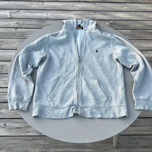 Grå hoodie från Polo Ralph Lauren - Säljer en klassisk grå hoodie från Polo Ralph Lauren med dragkedja och huva. Tröjan har två fickor framtill och den ikoniska broderade loggan på bröstet. Perfekt för en avslappnad stil. Nypris 1200 mitt pris 499kr. Pris går att diskutera. 