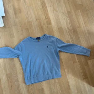 Ljusblå tröja från Polo Ralph Lauren - Ljusblå tröja från Polo Ralph Lauren med rund halsringning och diskret broderad logga på bröstet. Tröjan har långa ärmar och är tillverkad i mjuk bomull, perfekt för en avslappnad stil.