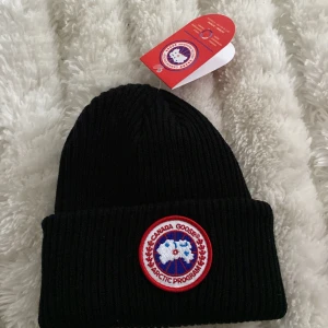 Svart ribbad mössa från Canada Goose - Svart ribbstickad mössa från Canada Goose med klassisk rund broderad logotyp framtill. Mössan är i tjockt material och har en uppvikt kant. Perfekt för kalla dagar.