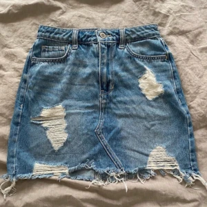 Blå jeanskjol med slitningar från Hollister - Säljer en blå jeanskjol från Hollister med flera slitna detaljer och fransig nederkant. Står storlek 0, passar som XS/S. 