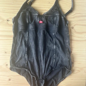 Svart body från Diesel - Säljer en svart body från Diesel med coola sömdetaljer och bred resår under bysten. Bodyn har justerbara axelband och en sportig look med en liten röd Diesel-logga framtill. Strl S