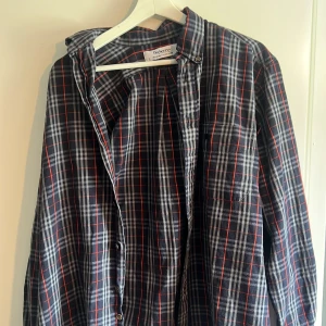 Rutig skjorta från Burberrys - Säljer en klassisk rutig skjorta från Burberrys i mörkblått, vitt och rött. Skjortan har långa ärmar, knappar framtill och vid ärmslut samt en bröstficka. Perfekt för dig som gillar tidlös stil och mönster.