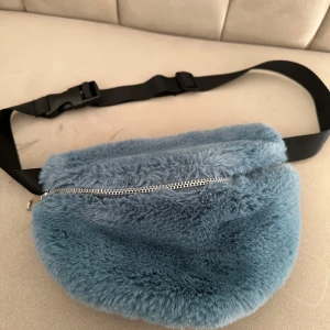 Blå fluffig magväska med dragkedja - Supermjuk och fluffig blå magväska med silvrig dragkedja och justerbart svart band. Perfekt för att bära det viktigaste på ett trendigt sätt. Väskan har en modern look och känns riktigt mysig.
