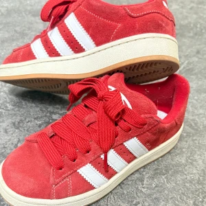 Röda Adidas Campus sneakers - Säljer ett par röda Adidas Campus sneakers i mocka med vita klassiska ränder och vit sula. Skorna har breda röda skosnören och låg profil. Perfekta för dig som gillar sportig och retro stil. 