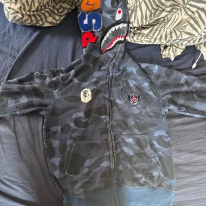 Camouflage zip-hoodie från A Bathing Ape - Säljer en mörkblå och grå camouflage hoodie från A Bathing Ape med hajmotiv på huvan, broderade bokstäver och tigerdetalj. Jackan har dragkedja, fickor fram och ribbade muddar. Perfekt för dig som gillar streetwear och unika plagg.