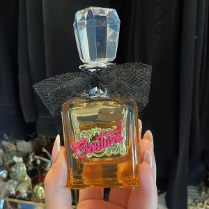 Viva La Juicy Gold Couture Eau de Parfum - En lyxig parfymflaska i glas med guldtonad vätska, dekorerad med svart tyllrosett och en stor, kristalliknande kork. Framsidan har en guldfärgad detalj med rosa texten 'Couture'. Doften är Eau de Parfum.