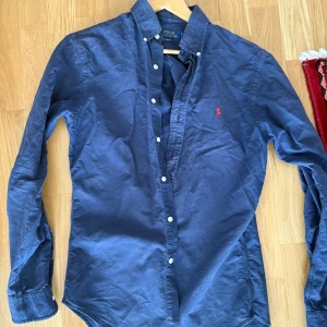 Mörkblå skjorta från Ralph Lauren - Säljer en klassisk mörkblå skjorta från Ralph Lauren med röd broderad logga på bröstet. Skjortan har knappar hela vägen, button-down krage och långa ärmar. Perfekt till jeans eller chinos. Har blivit för liten för mig och därför är passformen inte så bra!