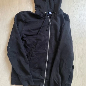 Svart hoodie med dragkedja från Divided - Svart hoodie från Divided med dragkedja framtill och huva med dragsko. Tröjan har två fickor på magen och är tillverkad i mjukt material, perfekt för en avslappnad stil. Pyttelite nopprig på insidan