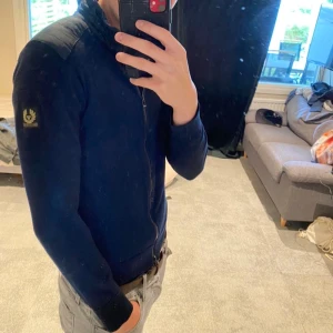 Mörkblå cardigan från Belstaff - Säljer en mörkblå cardigan från Belstaff med quiltade axelpartier. Tröjan har dragkedja hela vägen och ribbade muddar. Klassisk och stilren design, perfekt för lager-på-lager.