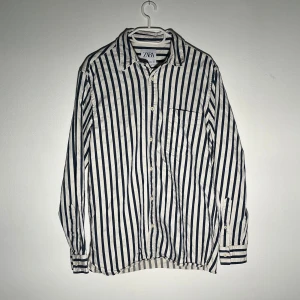 Randig skjorta från Zara - Säljer en vit skjorta med blåa vertikala ränder från Zara. Skjortan har klassisk krage, bröstficka och knäppning framtill. Perfekt för dig som gillar en stilren och avslappnad look.