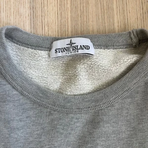 Grå sweatshirt från Stone Island Junior - Säljer en stilren grå sweatshirt från Stone Island Junior med klassisk rund halsringning och ikonisk logotyp-patch på ärmen. Tröjan har långa ärmar och är tillverkad i mjuk bomull för maximal komfort.