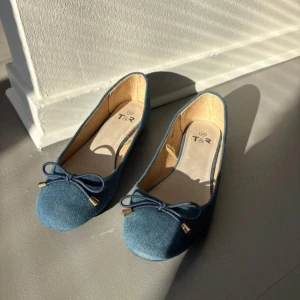 Blå ballerinaskor med rosett från T&R - Säljer ett par blå ballerinaskor från T&R med söt rosettdetalj framtill och små guldfärgade metalldetaljer. Skorna har rund tå och är tillverkade i ett tygmaterial som påminner om jeans.